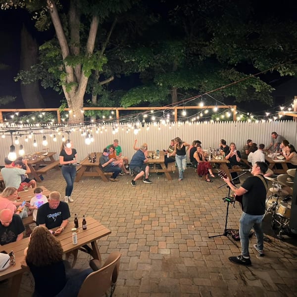 Night view of The Pieman Pizza's outdoor patio with string lights, diners, and a live musician.