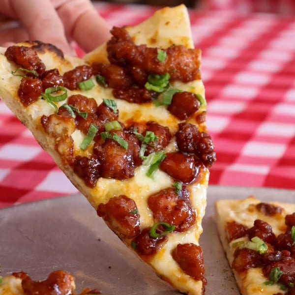 A hand holds up a slice of The Pieman Pizza's chicken pizza, with a red and white checkered tablecloth in the background.