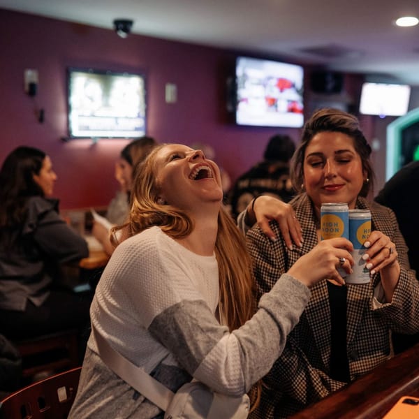Two women laughing and holding canned drinks at a bar in The Pieman Pizza.