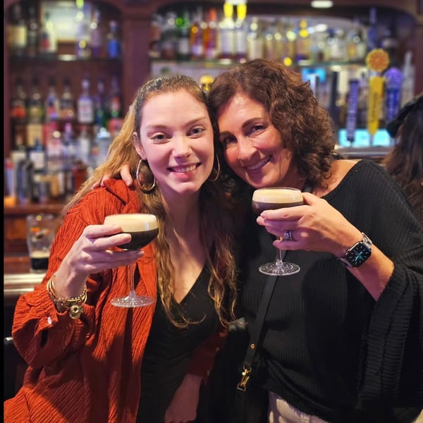 Two smiling women holding espresso martini glasses in front of a bar with liquor bottles.