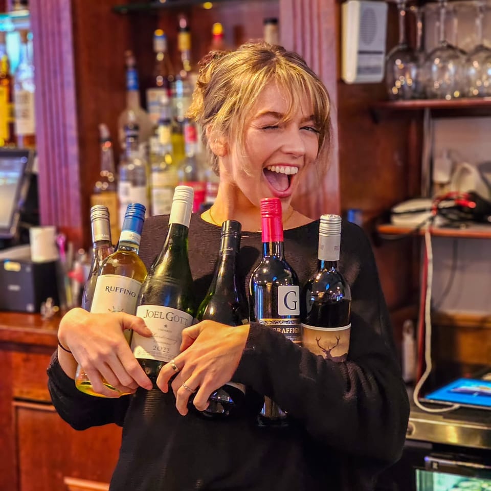 A smiling woman winks while holding five bottles of wine in a bar setting.