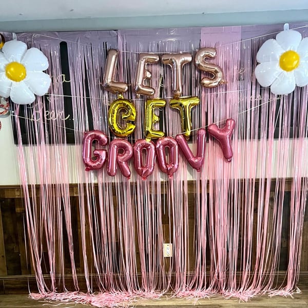 A pink fringe backdrop with "LET'S GET GROOVY" balloons and two white daisy balloons.