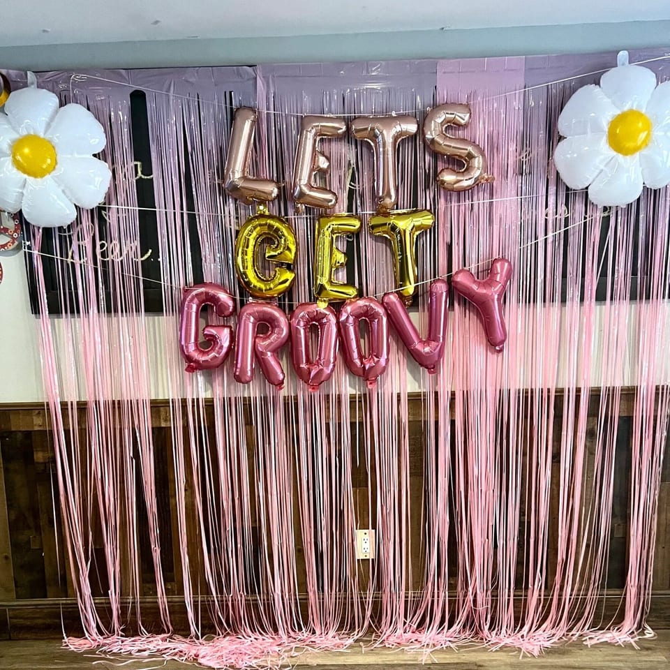 A pink fringe backdrop with "LET'S GET GROOVY" balloons and two white daisy balloons.