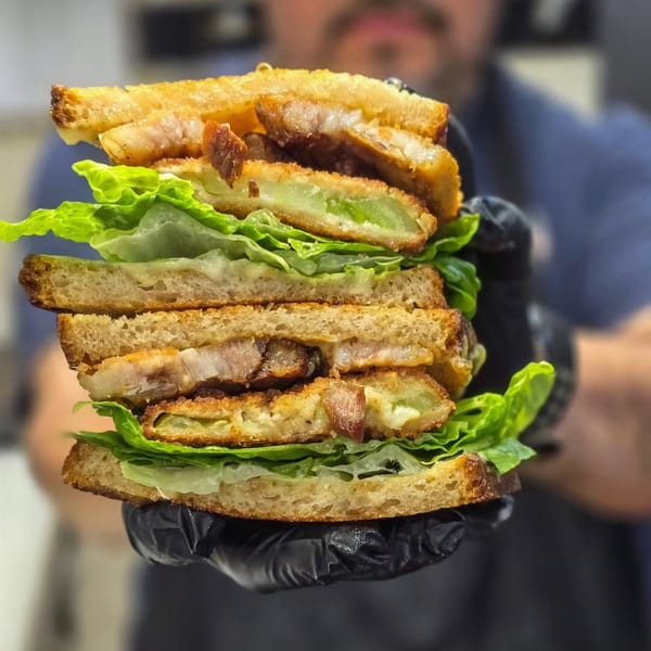 A large, stacked sandwich with crispy fried green tomatoes, bacon, and lettuce, held by gloved hands.