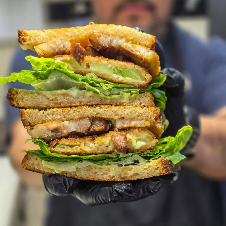 A large, stacked sandwich with crispy fried green tomatoes, bacon, and lettuce, held by gloved hands.