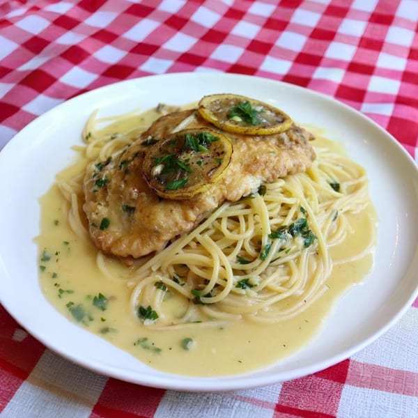 Chicken piccata with spaghetti, lemon slices, and parsley on a white plate over a red and white checkered tablecloth.
