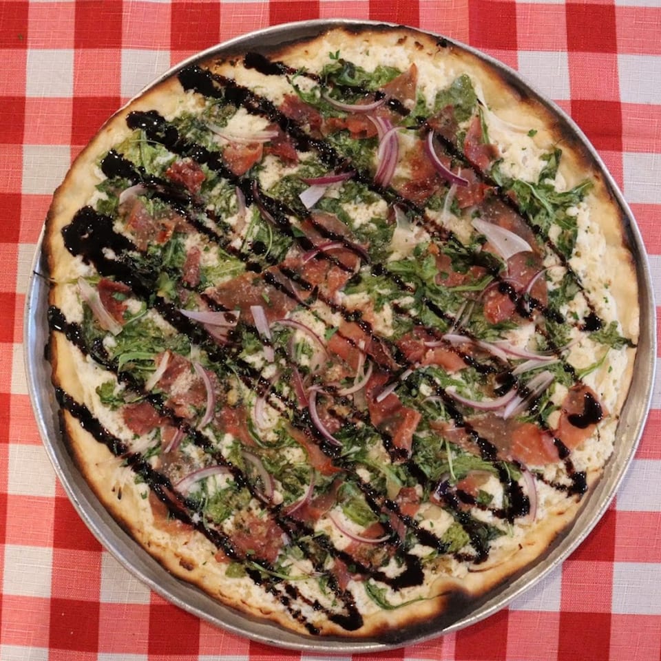 The Pieman Pizza's white pizza with arugula, prosciutto, red onion, and balsamic glaze on a red checkered tablecloth.