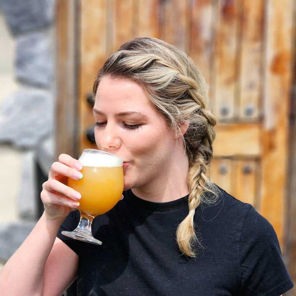 A woman with braided blonde hair sips a frothy orange drink from a stemmed glass.